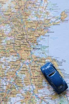 A detailed map of the Boston area with a small blue toy car placed on top of it. The map shows various cities and towns, highlighted roads and highways, and geographic features in the northeastern United States.