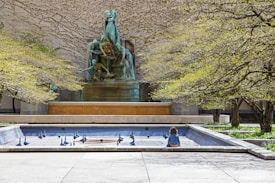 A young child with curly hair sits alone by a dry, rectangular fountain surrounded by sculptural installations. In the background, a large, ornate statue features multiple human figures made of what appears to be bronze. The scene is framed by trees with budding leaves, suggesting early spring.