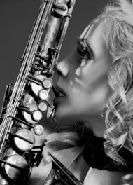 A close-up, black-and-white image of a woman with curly hair holding a saxophone. The focus is on her profile and the intricate details of the saxophone, with shadows creating a dramatic effect on her face.