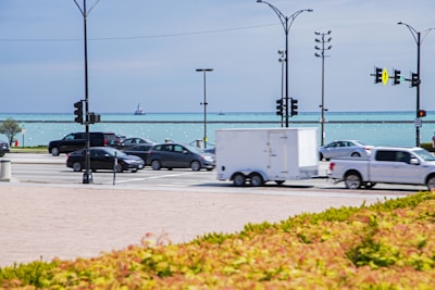 A busy street scene features several vehicles, including cars and trucks, driving along a road parallel to a vast body of water. Traffic lights and street lamps are visible, and a small boat is seen in the distance on the water.