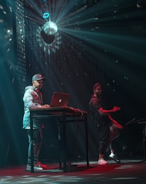 A DJ is performing on stage with a laptop placed on a high, narrow table. The dimly-lit environment is enhanced by a disco ball above, casting light beams around the room. Another person stands on the right, enthusiastically playing a drum.