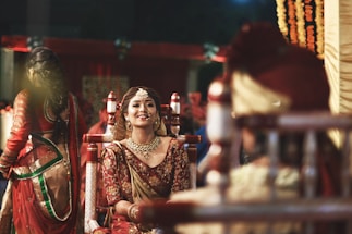 A bride dressed in traditional Indian wedding attire is smiling, adorned with intricate jewelry and a maang tikka, against a vibrant background. Other people in ceremonial clothing are present, contributing to a festive and cultural atmosphere.