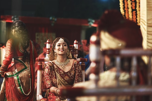 A bride dressed in traditional Indian wedding attire is smiling, adorned with intricate jewelry and a maang tikka, against a vibrant background. Other people in ceremonial clothing are present, contributing to a festive and cultural atmosphere.