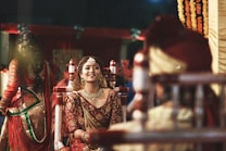 A bride dressed in traditional Indian wedding attire is smiling, adorned with intricate jewelry and a maang tikka, against a vibrant background. Other people in ceremonial clothing are present, contributing to a festive and cultural atmosphere.