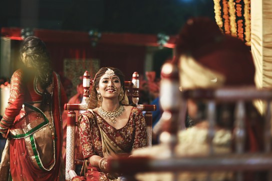 A bride dressed in traditional Indian wedding attire is smiling, adorned with intricate jewelry and a maang tikka, against a vibrant background. Other people in ceremonial clothing are present, contributing to a festive and cultural atmosphere.