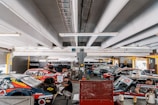 A busy garage area featuring several race cars with vivid decals and sponsorships. Mechanics and crew members work around the vehicles, indicating preparation or maintenance activities. The ceiling is lined with fluorescent lights, and various automotive equipment and tools are visible in the background. The scene is set in an industrial, utilitarian space, with a focus on motorsport.