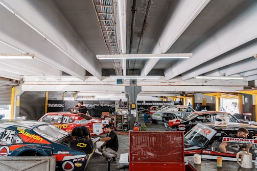 A busy garage area featuring several race cars with vivid decals and sponsorships. Mechanics and crew members work around the vehicles, indicating preparation or maintenance activities. The ceiling is lined with fluorescent lights, and various automotive equipment and tools are visible in the background. The scene is set in an industrial, utilitarian space, with a focus on motorsport.