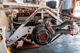 A partially disassembled vehicle engine with various mechanical components and wiring visible. The engine appears to be mounted on a frame, with a brake disc and caliper in the forefront. There's a person standing in the background, partially visible from the waist down, observing the scene.
