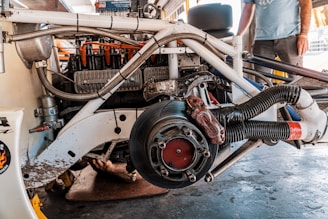 A partially disassembled vehicle engine with various mechanical components and wiring visible. The engine appears to be mounted on a frame, with a brake disc and caliper in the forefront. There's a person standing in the background, partially visible from the waist down, observing the scene.