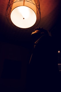 A person is looking up towards a ceiling light fixture that is casting a warm glow in a dark room. The silhouette of the person is visible against the bright light, creating a strong contrast.