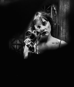 A person is holding a vintage camera close to their face. The image is in black and white, with focused lighting on the person’s face, highlighting delicate facial details and tattoos on the shoulder. The background appears dimly lit, creating a dramatic contrast.