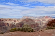 A vast and majestic canyon landscape with layers of red and beige rock formations stretching into the distance. The sky above is partly cloudy with patches of blue. Sparse vegetation is visible in the foreground, providing contrast to the rugged terrain.