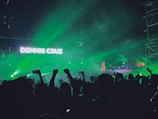 A vibrant concert scene with green lights illuminating a crowd of people raising their hands and holding phones. A large screen displays the name 'Dennis Cruz' with stage equipment visible in the background.