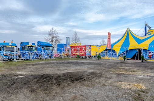 A vibrant circus scene with a large striped yellow and blue tent. In front of the tent, several brightly colored trucks are parked, featuring bold and playful designs, alongside banners and fencing. Sparse trees and a cloudy sky form the backdrop.