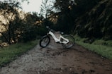 A white electric bike is parked on a muddy, forested trail. The path is surrounded by lush green vegetation and trees, with a peaceful, natural ambiance. The bike is positioned at an angle, highlighting its modern design and thick tires, suitable for off-road journeys.