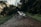 A white electric bike is parked on a muddy, forested trail. The path is surrounded by lush green vegetation and trees, with a peaceful, natural ambiance. The bike is positioned at an angle, highlighting its modern design and thick tires, suitable for off-road journeys.