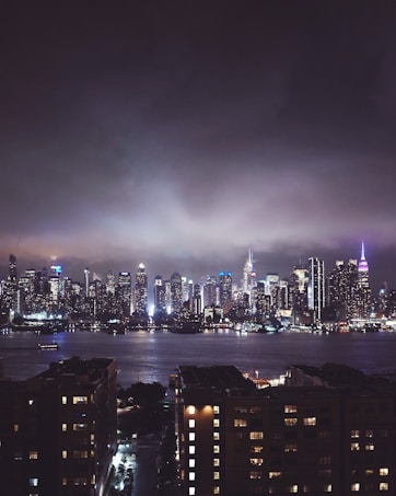 A nighttime cityscape with a brightly lit skyline across a body of water. The sky is cloudy, adding a dramatic effect to the scene. The buildings are illuminated, creating a vibrant glow against the dark sky. In the foreground, there are lower buildings and streets with dimmer lighting.