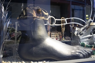 A large, metallic, sculpture of a foot is displayed in a storefront window. The foot is set atop a bed of small, smooth stones. Around the foot, there's an assortment of items, including chains, a hat, and decorative pieces that resemble store merchandise.