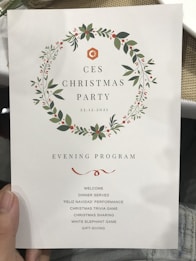 A program menu for the CES Christmas Party held on December 23, 2021. The design is adorned with a wreath made of green leaves and red berries. The evening program includes welcome, dinner, a performance titled 'Feliz Navidad', a Christmas trivia game, sharing session, White Elephant game, and gift-giving.