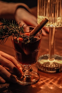 A warmly lit scene featuring a glass of dark beverage garnished with rosemary and a cinnamon stick. A hand reaches towards the glass, which is placed on a wooden table next to a decorative lamp. The light casts a soft glow, creating a cozy ambiance.