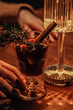 A warmly lit scene featuring a glass of dark beverage garnished with rosemary and a cinnamon stick. A hand reaches towards the glass, which is placed on a wooden table next to a decorative lamp. The light casts a soft glow, creating a cozy ambiance.
