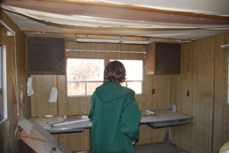 A person in a green coat is standing in a dilapidated room with wooden paneled walls and a partially broken ceiling. The room is cluttered, with scattered papers, a can, and debris on a desk-like surface. There are small cupboards above the windows, and the space looks abandoned.