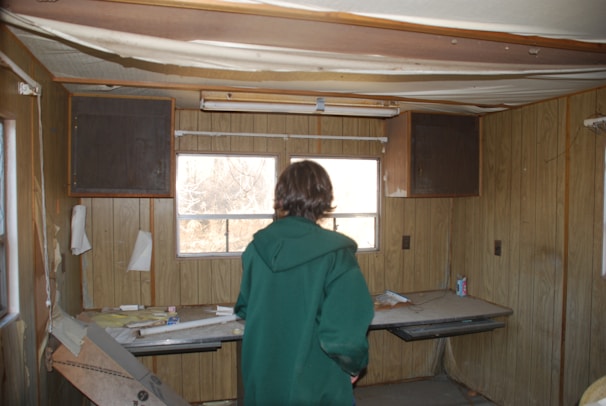 A person in a green coat is standing in a dilapidated room with wooden paneled walls and a partially broken ceiling. The room is cluttered, with scattered papers, a can, and debris on a desk-like surface. There are small cupboards above the windows, and the space looks abandoned.