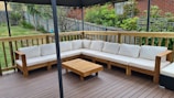 An outdoor patio features a large L-shaped wooden sofa with white cushions, set on a wooden deck. A small wooden coffee table is placed in front of the sofa. The area is surrounded by a wooden fence with a garden full of greenery and plants in the background.
