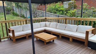 An outdoor patio features a large L-shaped wooden sofa with white cushions, set on a wooden deck. A small wooden coffee table is placed in front of the sofa. The area is surrounded by a wooden fence with a garden full of greenery and plants in the background.