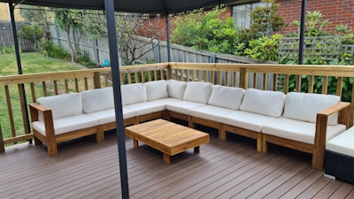 An outdoor patio features a large L-shaped wooden sofa with white cushions, set on a wooden deck. A small wooden coffee table is placed in front of the sofa. The area is surrounded by a wooden fence with a garden full of greenery and plants in the background.