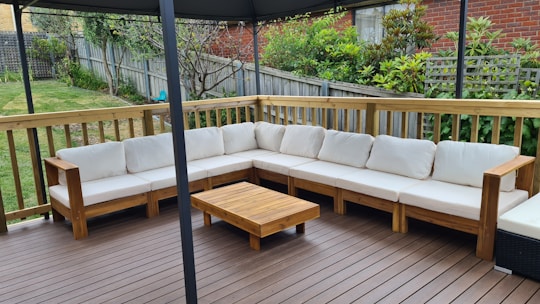 An outdoor patio features a large L-shaped wooden sofa with white cushions, set on a wooden deck. A small wooden coffee table is placed in front of the sofa. The area is surrounded by a wooden fence with a garden full of greenery and plants in the background.