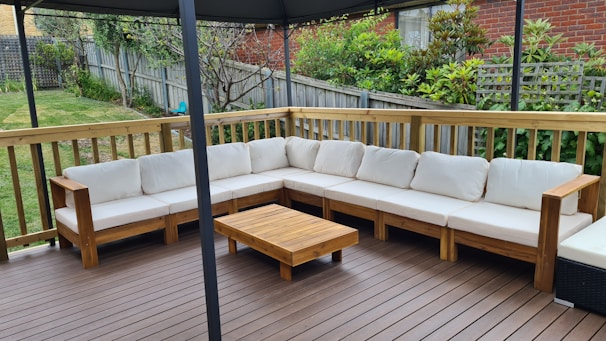 An outdoor patio features a large L-shaped wooden sofa with white cushions, set on a wooden deck. A small wooden coffee table is placed in front of the sofa. The area is surrounded by a wooden fence with a garden full of greenery and plants in the background.