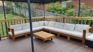 An outdoor patio features a large L-shaped wooden sofa with white cushions, set on a wooden deck. A small wooden coffee table is placed in front of the sofa. The area is surrounded by a wooden fence with a garden full of greenery and plants in the background.