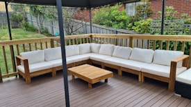 An outdoor patio features a large L-shaped wooden sofa with white cushions, set on a wooden deck. A small wooden coffee table is placed in front of the sofa. The area is surrounded by a wooden fence with a garden full of greenery and plants in the background.