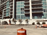 A modern multi-story building with numerous glass balconies stands in the background. In the foreground, there are several traffic cones and barriers on a rough, unfinished surface, indicating a construction or road work site. Palm trees are planted along a white wall in front of the building, adding a touch of greenery.