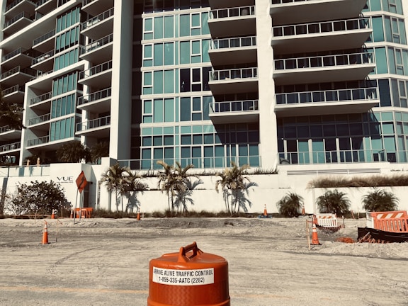 A modern multi-story building with numerous glass balconies stands in the background. In the foreground, there are several traffic cones and barriers on a rough, unfinished surface, indicating a construction or road work site. Palm trees are planted along a white wall in front of the building, adding a touch of greenery.