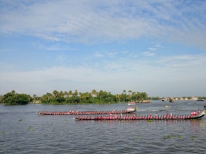 Two long boats with a large number of rowers dressed in pink are racing on a wide river. The background features lush green trees and palm trees along the riverbank, under a partly cloudy blue sky. Some small boats and a houseboat can be seen in the distance.