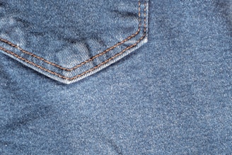 A close-up view of denim fabric featuring a corner of a pocket with detailed orange stitching.