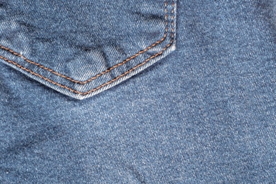A close-up view of denim fabric featuring a corner of a pocket with detailed orange stitching.