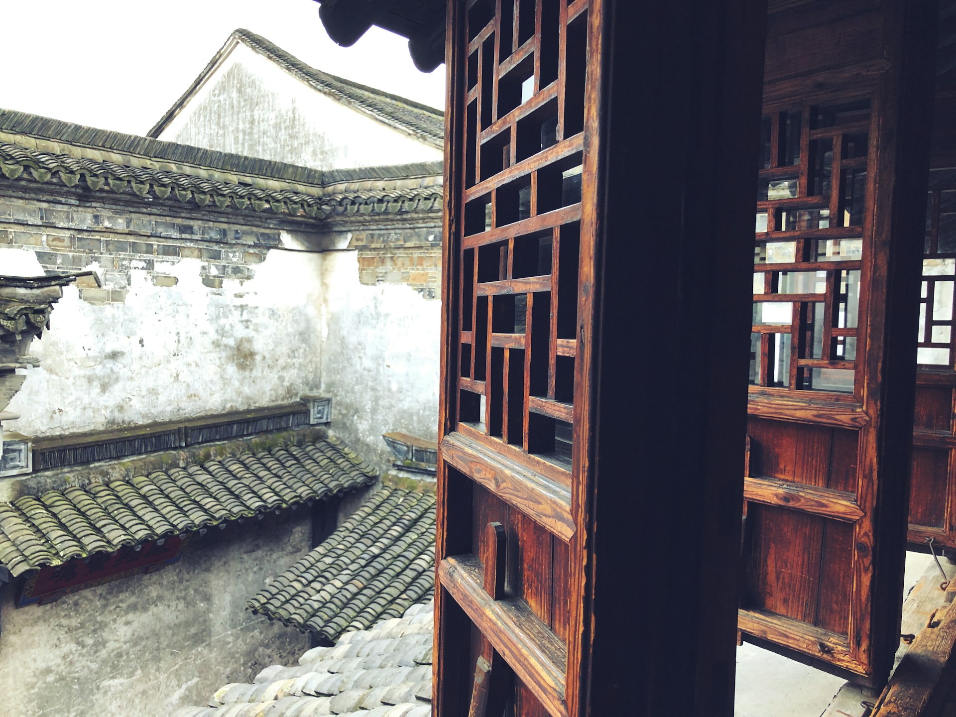 A traditional architectural structure features wooden lattice windows and tiled roofs. The building appears aged with weathered surfaces, showcasing intricate wooden carvings set against the backdrop of a courtyard with gray and mossy tiles.