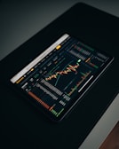 A tablet displaying a detailed digital financial trading interface with various graphs, charts, and numerical data. The screen shows candlestick charts with green and red indicators, illustrating market trends and stock prices.