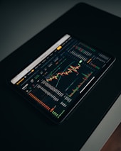 A tablet displaying a detailed digital financial trading interface with various graphs, charts, and numerical data. The screen shows candlestick charts with green and red indicators, illustrating market trends and stock prices.