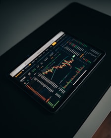 A tablet displaying a detailed digital financial trading interface with various graphs, charts, and numerical data. The screen shows candlestick charts with green and red indicators, illustrating market trends and stock prices.
