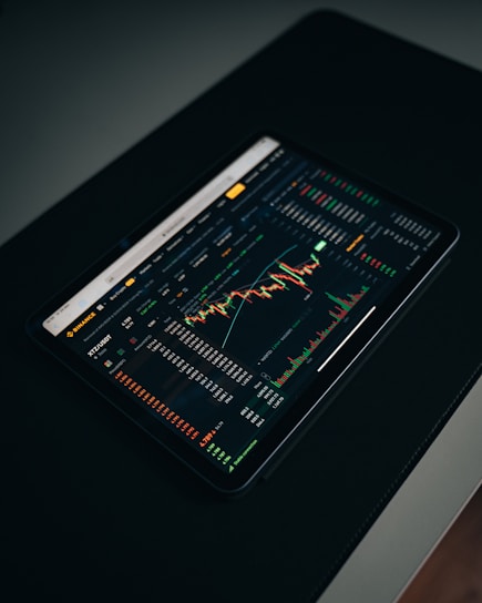 A tablet displaying a detailed digital financial trading interface with various graphs, charts, and numerical data. The screen shows candlestick charts with green and red indicators, illustrating market trends and stock prices.