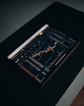 A tablet displaying a detailed digital financial trading interface with various graphs, charts, and numerical data. The screen shows candlestick charts with green and red indicators, illustrating market trends and stock prices.