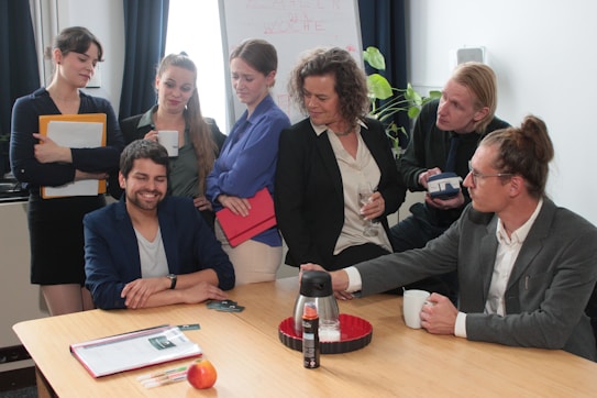 A group of seven people gathered around a conference table in an office environment. They appear to be engaged in a conversation, with one individual sitting at the table and others standing or leaning in. Various items such as folders, a thermos, a tray, and an apple are placed on the table. A flip chart and a plant are visible in the background.