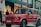 A bright red luxury SUV is parked on a city street in front of a Louis Vuitton store. Several people are standing nearby, dressed in winter clothing, suggesting cooler weather. The scene has a high-end, urban atmosphere with a mix of pedestrians and high-end stores.