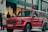 A bright red luxury SUV is parked on a city street in front of a Louis Vuitton store. Several people are standing nearby, dressed in winter clothing, suggesting cooler weather. The scene has a high-end, urban atmosphere with a mix of pedestrians and high-end stores.