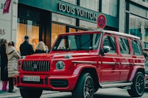 A bright red luxury SUV is parked on a city street in front of a Louis Vuitton store. Several people are standing nearby, dressed in winter clothing, suggesting cooler weather. The scene has a high-end, urban atmosphere with a mix of pedestrians and high-end stores.