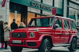 A bright red luxury SUV is parked on a city street in front of a Louis Vuitton store. Several people are standing nearby, dressed in winter clothing, suggesting cooler weather. The scene has a high-end, urban atmosphere with a mix of pedestrians and high-end stores.